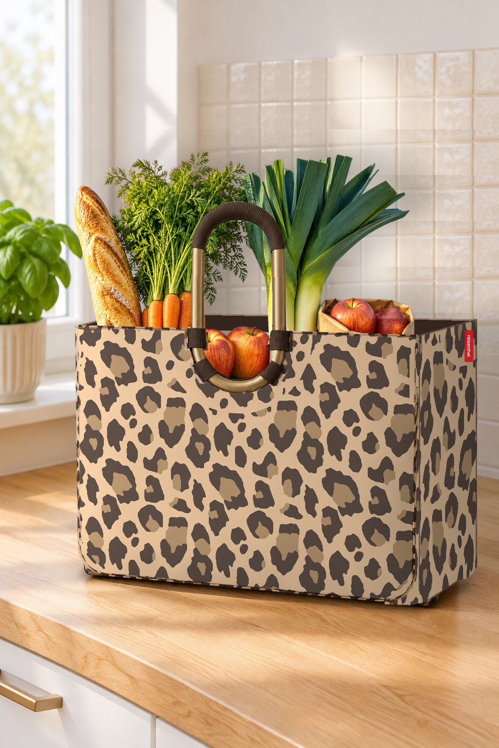 reisenthel loopshopper L frame im Leo-Macchiato-Muster leicht schräg auf einer hellen Eichen-Arbeitsplatte, gefüllt mit Baguette, Möhren mit Grün, Lauch und roten Äpfeln, rechts die Seitenwand mit rotem reisenthel-Etikett, links Fenster mit warmem Licht und Basilikum