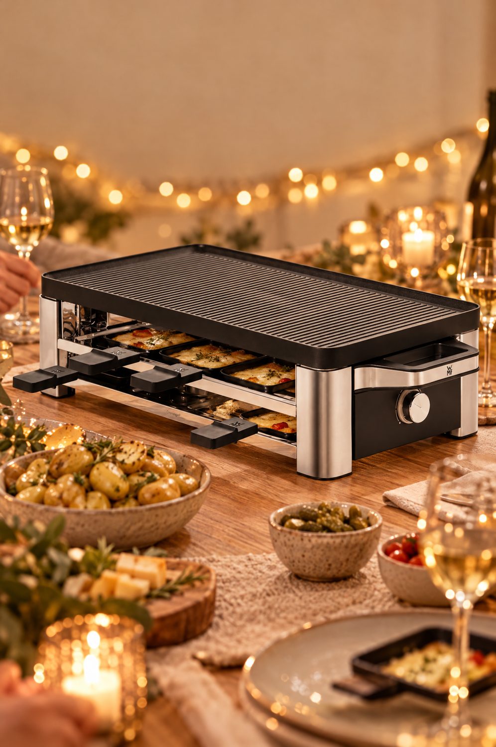 WMF Lono Raclette aus Edelstahl auf einem festlich gedeckten Tisch mit Pfännchen, Kartoffeln, Käse und Weingläsern, Lichterkette im Hintergrund