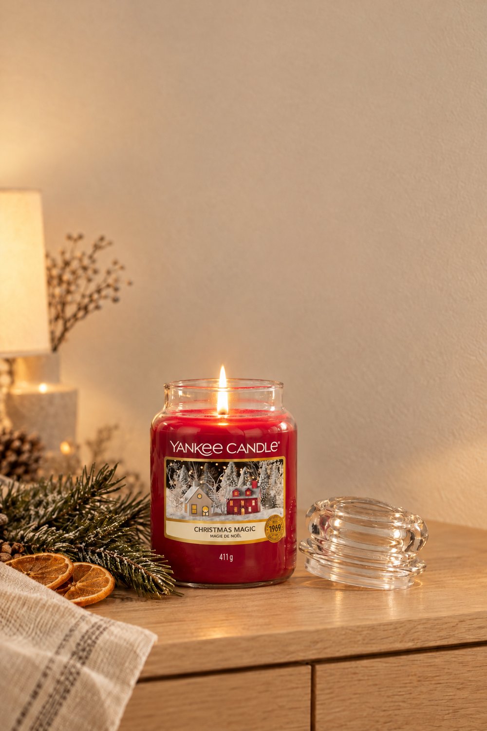 Rote Yankee Candle Christmas Magic im mittleren Glas brennt auf einem Holzsideboard neben Tannenzweig und getrockneten Orangenscheiben