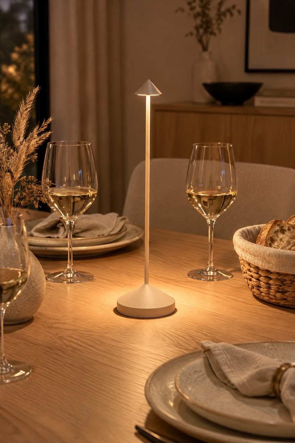 Eine einzelne schlanke Akku-Tischleuchte in Sand von Zafferano leuchtet warm in der Mitte eines gedeckten Holztischs zwischen Tellern und Weingläsern, Abendlicht im Esszimmer